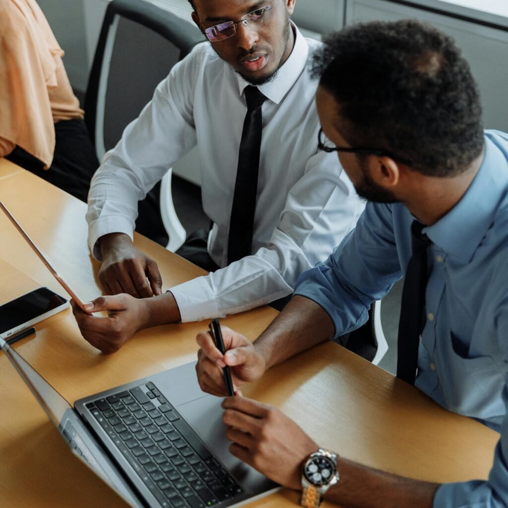 Two men in business attire discuss work at an office desk with a laptop.